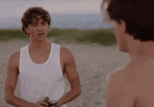 a man in a white tank top is standing next to another man without a shirt on the beach .