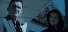a man in a suit stands next to another man in a hood