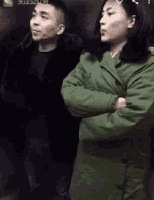 a man and a woman standing next to each other with their arms crossed . the woman is wearing a green coat .