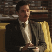 a man in a suit and tie is sitting on a yellow couch writing in a notebook .