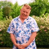 a man wearing a blue and white floral shirt is smiling in front of a bush with the words the great canadian baking show