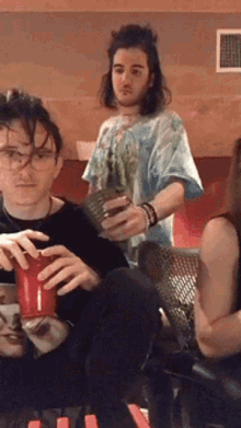 a group of people are sitting around a table with one man holding a red cup