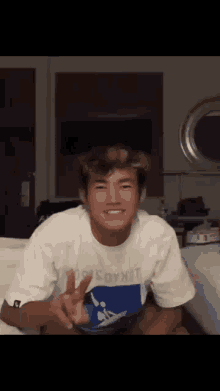 a man in a white shirt is sitting on a couch and giving a peace sign .