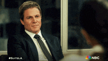 a man in a suit and tie is sitting in front of a window with a nbc logo on the bottom