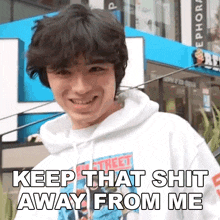 a young man wearing a white hoodie is smiling and says keep that shit away from me