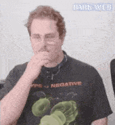 a man wearing glasses and a black t-shirt is covering his nose with his hand .