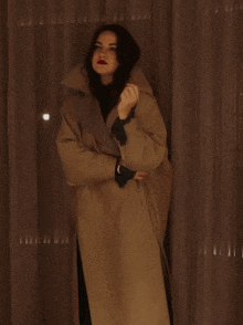 a woman in a tan coat is standing in front of a brown curtain