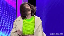 a woman in a trench coat and neon green sweater is dancing on a blue background .