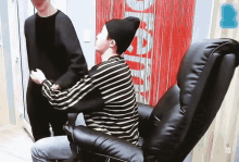 a man in a striped shirt sits in a chair in front of a red curtain with the word supreme on it