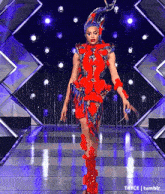 a drag queen is walking down a runway wearing a red and blue dress and a blue hat .