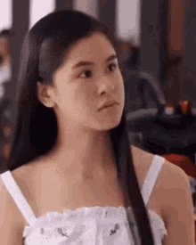 a woman with long black hair is wearing a white tank top and looking at the camera .