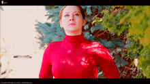 a woman in a red turtleneck stands in front of trees
