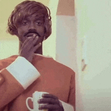 a man in a wig is holding a cup of coffee and covering his mouth .