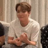 a man in a white t-shirt is sitting on a couch with his hands folded .