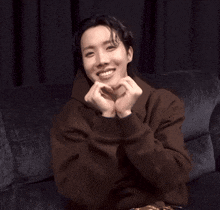 a young man in a brown sweatshirt is smiling and making a heart with his hands