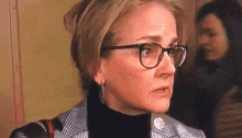 a woman wearing glasses and a black turtleneck is looking at the camera