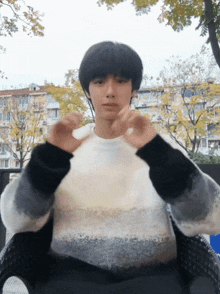 a young man in a sweater is sitting in a chair and making a heart shape with his hands
