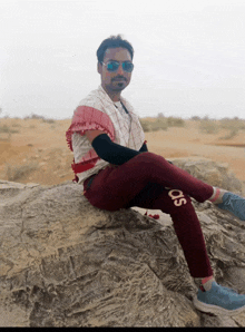a man wearing sunglasses sits on a rock with his legs crossed and his pants say so