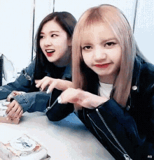 two girls are sitting at a table with a box of pizza . one of the girls is wearing a leather jacket .
