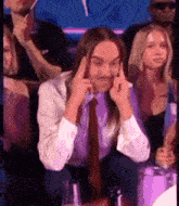 a man with long hair and a tie is sitting in a crowd of people and making a funny face .