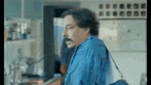 a man with a mustache and a blue shirt is walking in a kitchen .