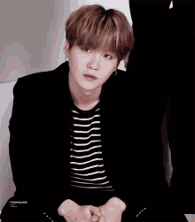 a young man wearing a black jacket and striped shirt is sitting down with his hands folded .