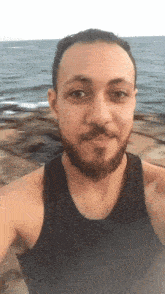a man with a beard wearing a black tank top is standing in front of the ocean