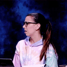 a woman wearing glasses and a tie dye sweatshirt is sitting in front of a laptop