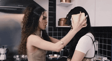 two women are standing in a kitchen with one holding a bread loaf over the other 's ear