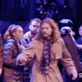 a man in a trench coat and plaid scarf stands in front of a group of people on a stage ..