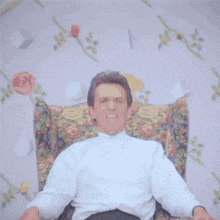 a man in a white shirt is sitting in a floral chair making a face .