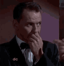 a man in a tuxedo and bow tie is covering his mouth with his hand .