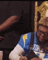 a man in a blue jersey is sitting in a chair writing on a piece of paper with a pen .