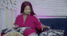 a woman in a pink jacket is sitting on a couch with pillows and a cell phone .