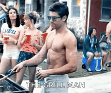 a shirtless man is cooking on a grill with a group of people behind him .