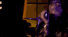 a woman is singing into a microphone while playing drums in a dark room with a window in the background .