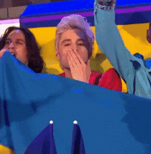 a man covering his mouth with his hand while sitting in front of a blue flag