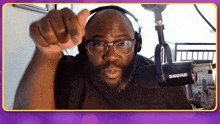 a man wearing glasses and headphones is pointing at the camera with a shure microphone in the background
