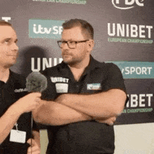 two men are standing next to each other with their arms crossed while a man is talking into a microphone .