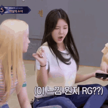 a girl in a white shirt is sitting on the floor and holding a cell phone while another girl looks on ..