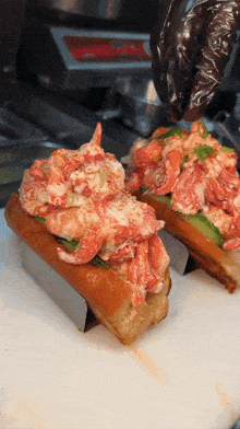 two lobster rolls are being prepared on a cutting board by a person wearing black gloves