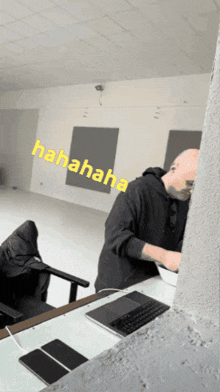 a man in a black hoodie sits at a desk with a laptop and a sign that says hahahaha