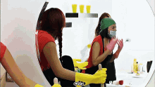 a woman wearing a red shirt and yellow gloves is cleaning a mirror