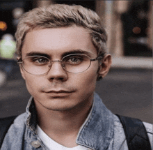 a young man wearing glasses and a denim jacket is looking at the camera