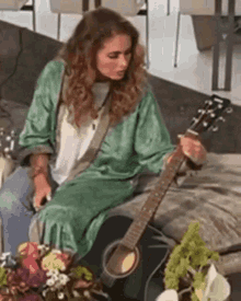 a woman in a green robe is playing an acoustic guitar while sitting on a couch .