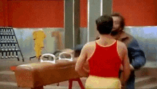 a man in a red tank top and yellow shorts is standing next to a man in a blue jacket .