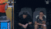 two men are sitting in a stadium watching a soccer game with the score at 1 to 1