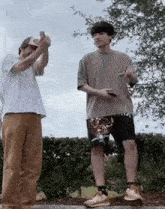 two young men are standing next to each other on a sidewalk . one of the men is taking a picture of the other .