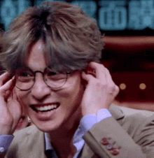 a young man wearing glasses and a suit is smiling and covering his ears .