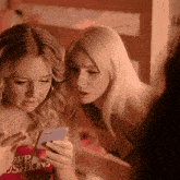 two women looking at a cell phone with one wearing a shirt that says " we cushions "
